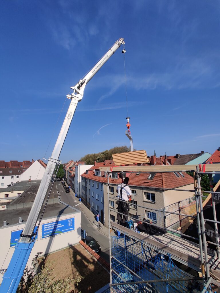 Ein Baukran hebt Materialien auf ein Dach, während ein Arbeiter auf einem Gerüst den Vorgang überwacht.