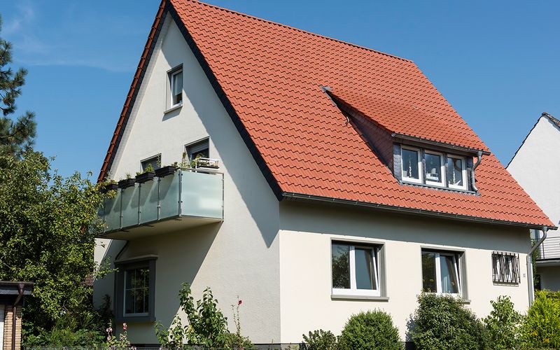 Ein Einfamilienhaus mit rotem Ziegeldach, helle Fassade, Balkon und Garten bei klarem Himmel.