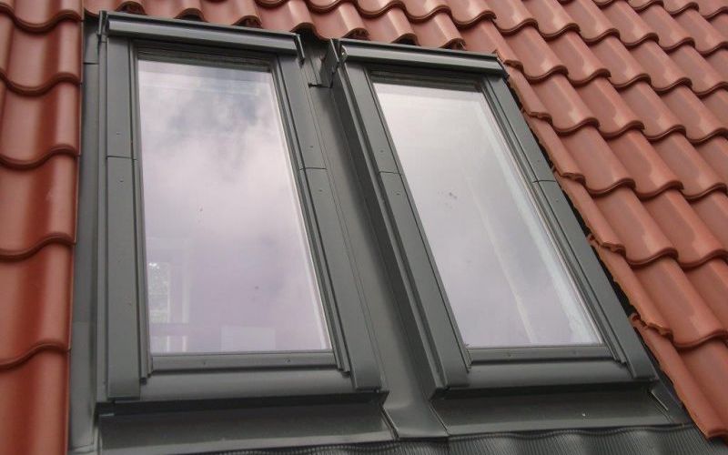 Zwei rechteckige Dachfenster in einem roten Ziegeldach, die den blauen Himmel reflektieren.