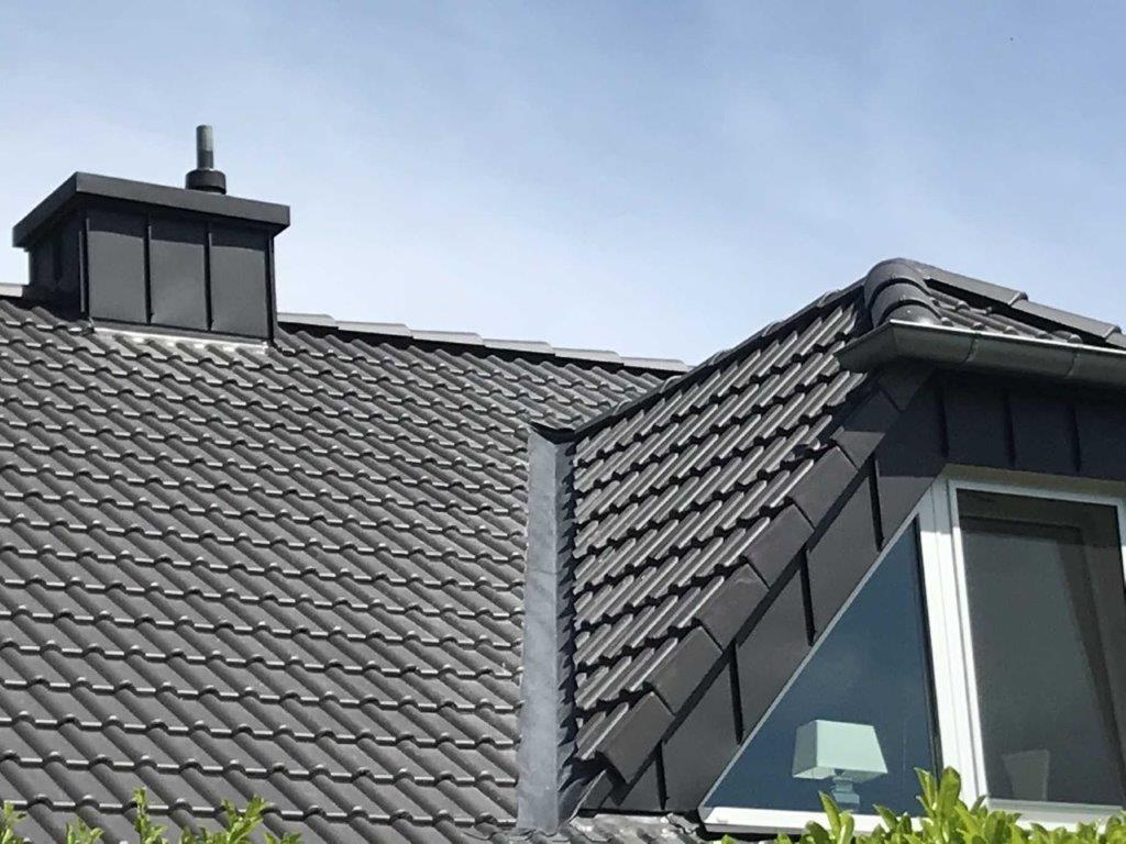 Ein modernes Hausdach mit dunkelgrauen Ziegeln, einer Gaube mit Fenster und einem kleinen Schornstein.