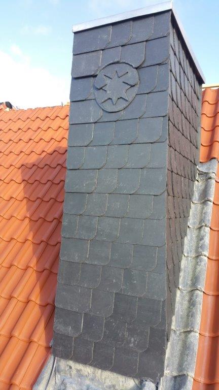 Ein Schornstein mit dunklen Schieferplatten auf einem roten Ziegeldach, verziert mit einem Stern.