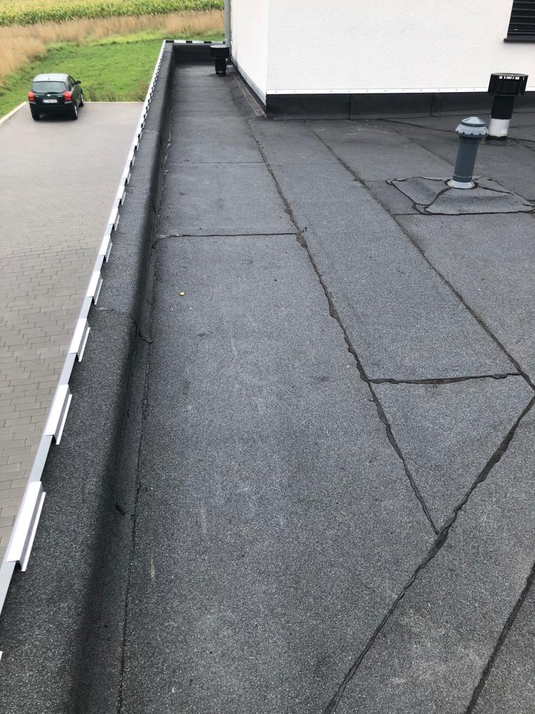 Ein Flachdach mit dunklem Bitumen, Lüftungsrohr und technischen Vorrichtungen, umgeben von grünem Gras.