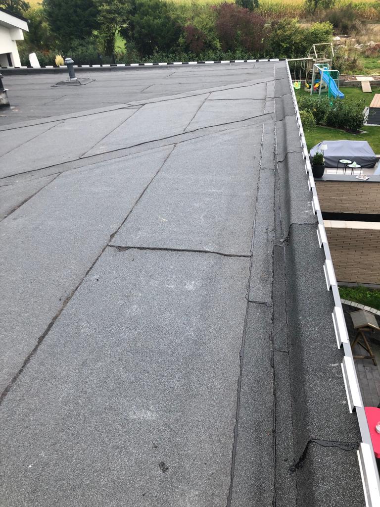 Ein Flachdach mit dunkler Dachpappe, umgeben von grüner Landschaft und einem Garten mit Spielgerät.