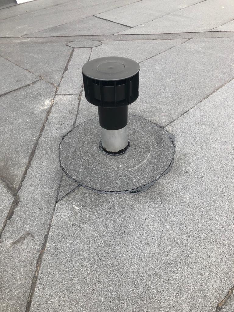 Ein silbernes Lüftungsrohr mit schwarzer Abdeckung ragt aus einer grauen, rauen Dachfläche heraus.