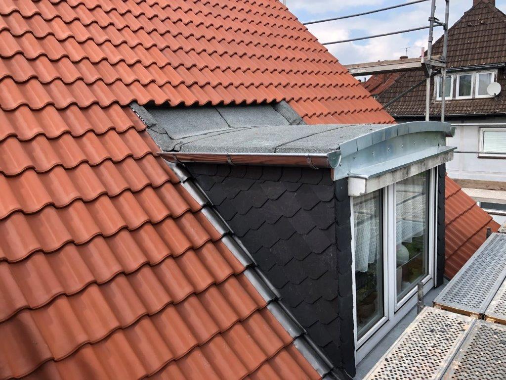 Ein Dach mit roten Ziegeln, einer Gaube aus dunklen Schieferplatten und einem weißen Dachfenster.
