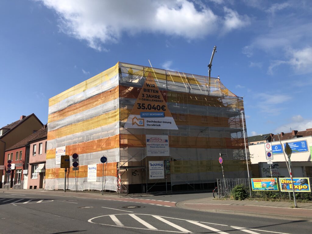 Ein Gebäude ist vollständig in ein orange-weißes Gerüstnetz gehüllt, mit einem Banner zur Ausbildung.