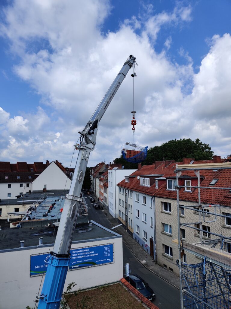 Ein blauer Kran hebt eine mit Netz gesicherte Last über Wohnhäuser mit roten Dächern in einer Stadt.