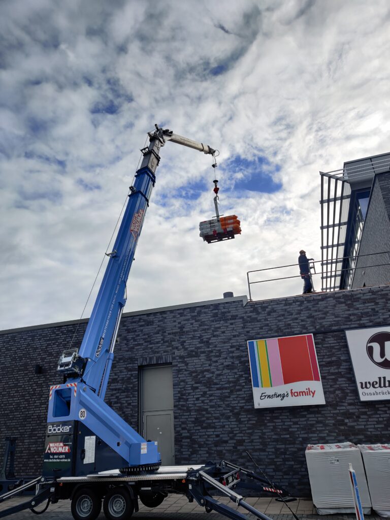 Ein blauer Kran hebt Baumaterialien auf das Dach eines Ziegelgebäudes, während eine Person zusieht.