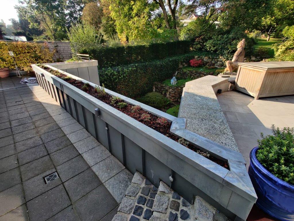 Ein gepflegter Garten mit Pflanzkästen, blauer Schale, Skulptur und Kletterpflanzen an der Mauer.
