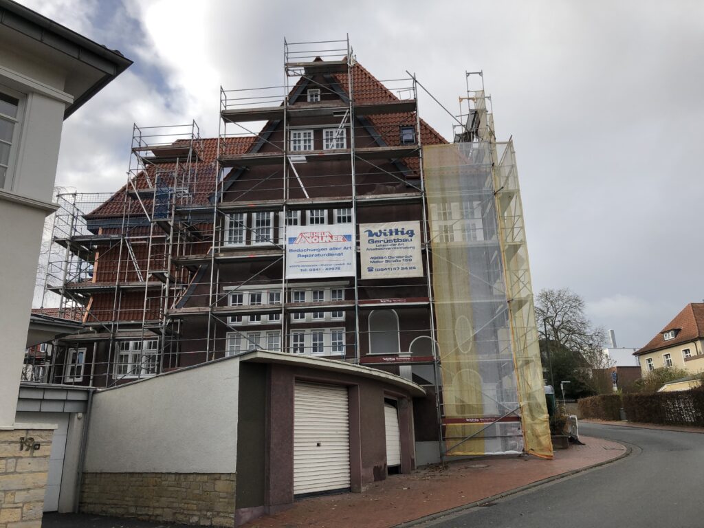 Ein mehrstöckiges Gebäude mit rotem Ziegeldach ist von einem silbernen Gerüst für Renovierungsarbeiten umgeben.