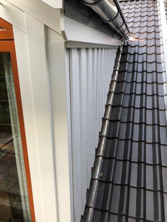 Dach mit dunkelgrauen Ziegeln, silberner Dachrinne, weißer Wand und Holzrahmenfenster.